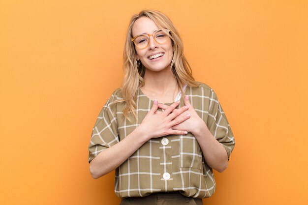 Young pretty blonde woman feeling romantic, happy and in love, smiling cheerfully and holding hands close to heart against flat color wall