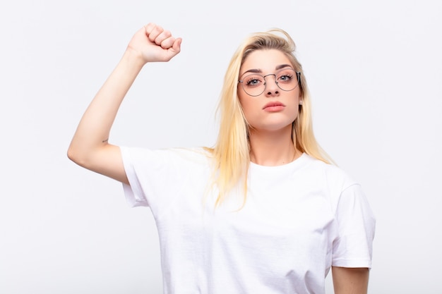 Giovane bella donna bionda sentendosi seria, forte e ribelle, alzando il pugno, protestando o combattendo per la rivoluzione contro il muro bianco