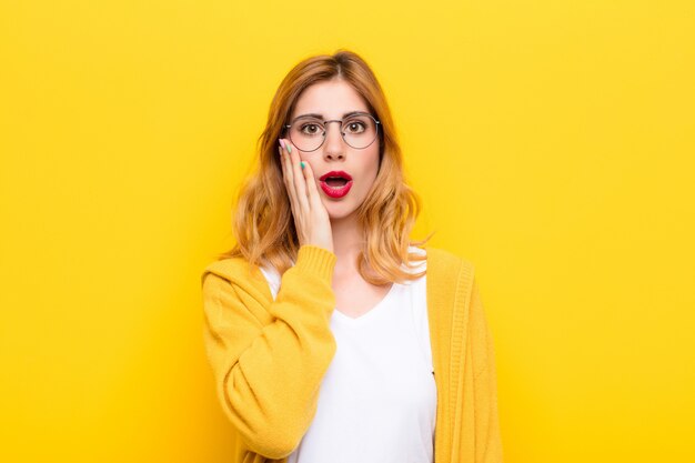 Young pretty blonde woman feeling shocked and astonished holding face to hand in disbelief with mouth wide open against yellow wall