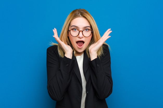 Young pretty blonde woman feeling shocked and excited, laughing, amazed and happy because of an unexpected surprise against flat wall