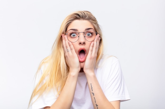 Young pretty blonde woman feeling shocked and scared, looking terrified with open mouth and hands on cheeks against white wall