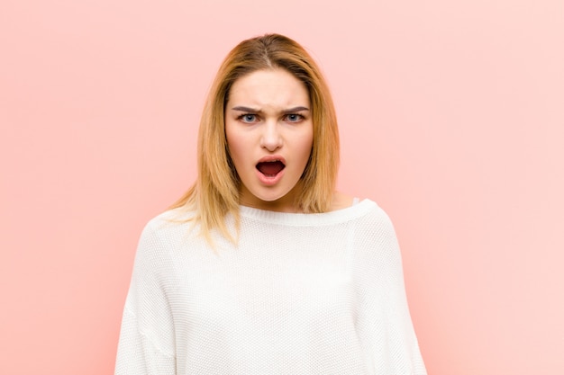 Young pretty blonde woman feeling terrified and shocked, with mouth wide open in surprise against pink flat wall