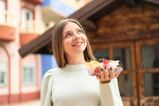 笑みを浮かべながら見上げて屋外でフルーツ ボウルを保持している若いきれいなブロンドの女性