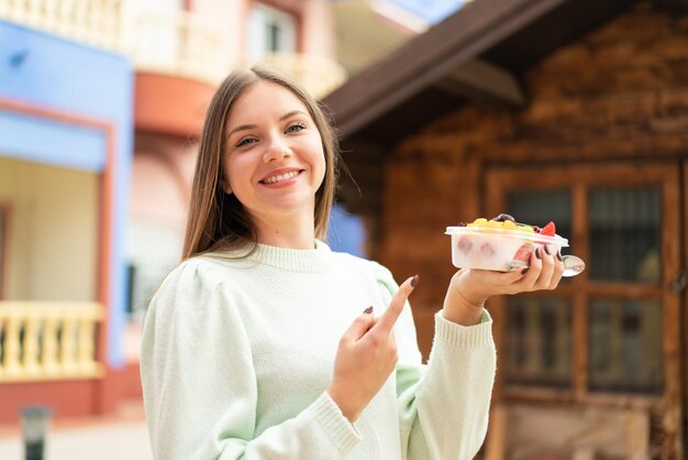 屋外で果物のボウルを保持し、それを指している若いきれいなブロンドの女性