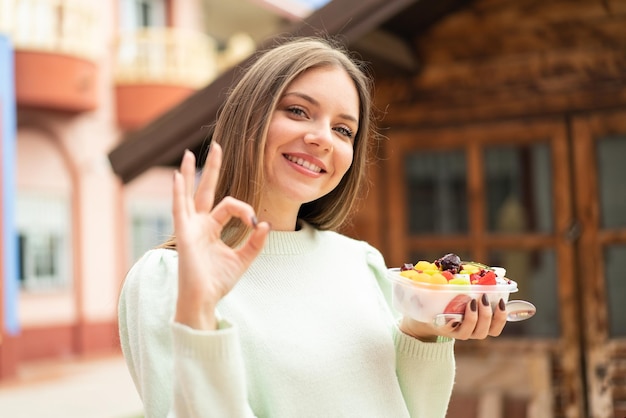 屋外でフルーツ ボウルを保持している若いきれいな金髪の女性指で ok のサインを示す