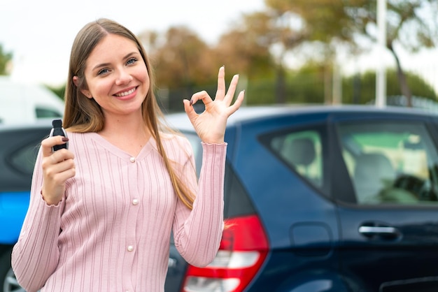 屋外で車のキーを保持している若いきれいなブロンドの女性指で ok のサインを示す