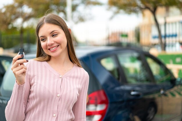 Молодая симпатичная блондинка держит ключи от машины на улице со счастливым выражением лица