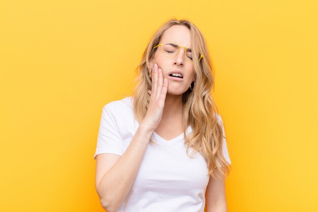 Young pretty blonde woman holding cheek and suffering painful toothache, feeling ill, miserable and unhappy, looking for a dentist against color wall