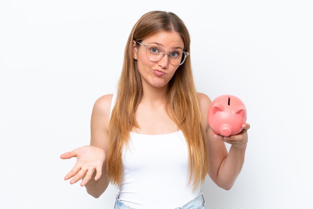 Young pretty blonde woman holding piggy bank isolated on white background making doubts gesture while lifting the shoulders