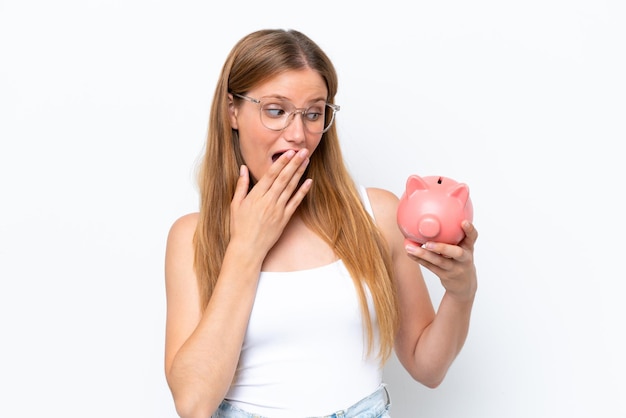 Young pretty blonde woman holding piggy bank isolated on white background with surprise and shocked facial expression