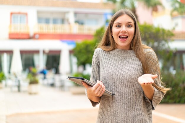 ショックを受けた表情で屋外でタブレットを保持している若いきれいなブロンドの女性