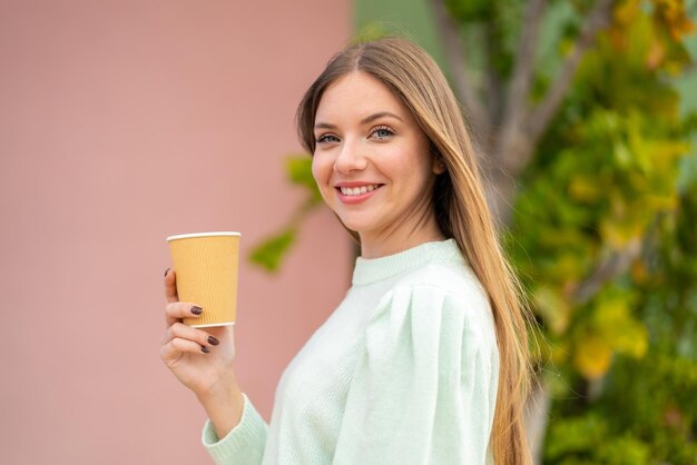 屋外でたくさんの笑顔でテイクアウト コーヒーを保持している若いきれいなブロンドの女性