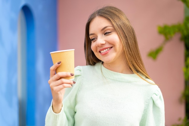 幸せな表情で屋外でテイクアウト コーヒーを保持している若いきれいなブロンドの女性