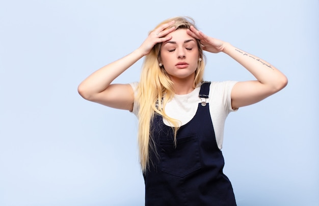 Young pretty blonde woman looking concentrated, thoughtful and inspired, brainstorming and imagining with hands on forehead against flat wall