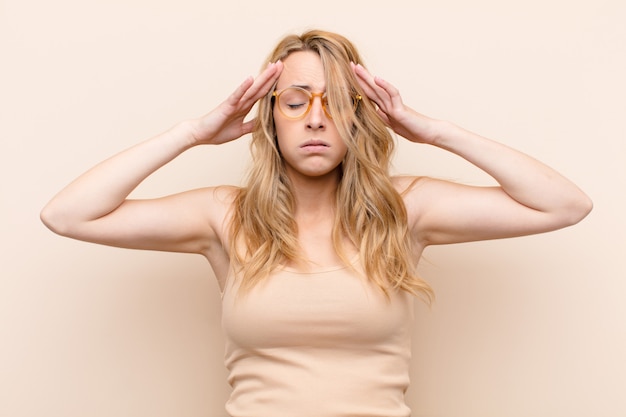 Young pretty blonde woman looking concentrated, thoughtful and inspired, brainstorming and imagining with hands on forehead over color wall