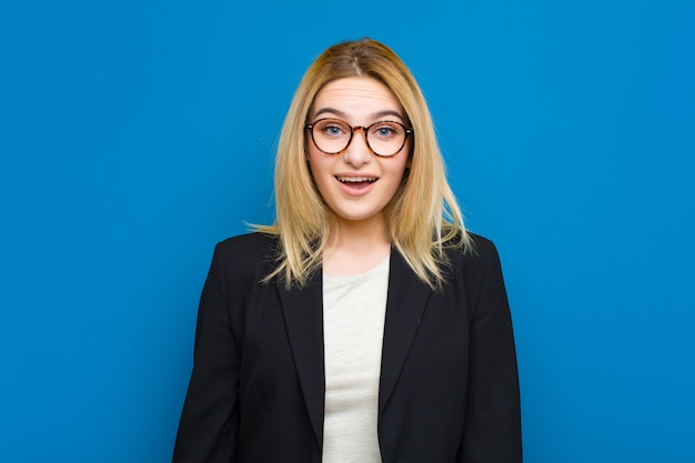 Young pretty blonde woman looking happy and pleasantly surprised, excited with a fascinated and shocked expression against flat wall