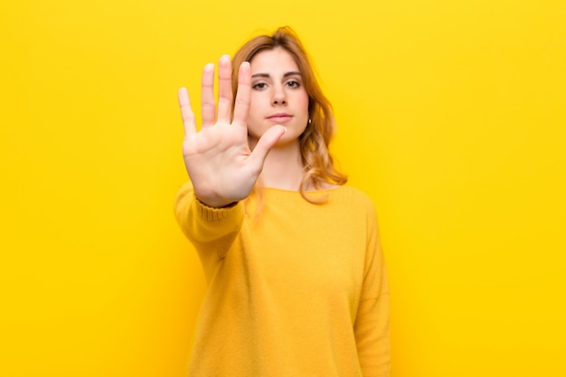 Young pretty blonde woman looking serious, stern, displeased and angry showing open palm making stop gesture against yellow wall