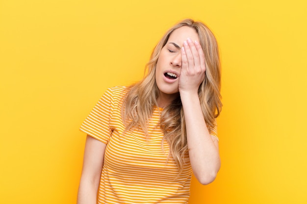 Young pretty blonde woman looking sleepy, bored and yawning, with a headache and one hand covering half the face against flat color wall