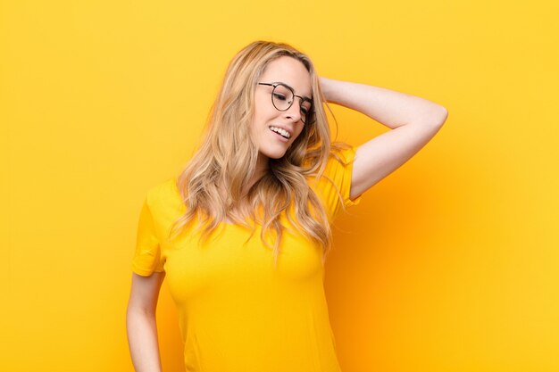 Young pretty blonde woman smiling cheerfully and casually, taking hand to head with a positive, happy and confident look against flat color wall