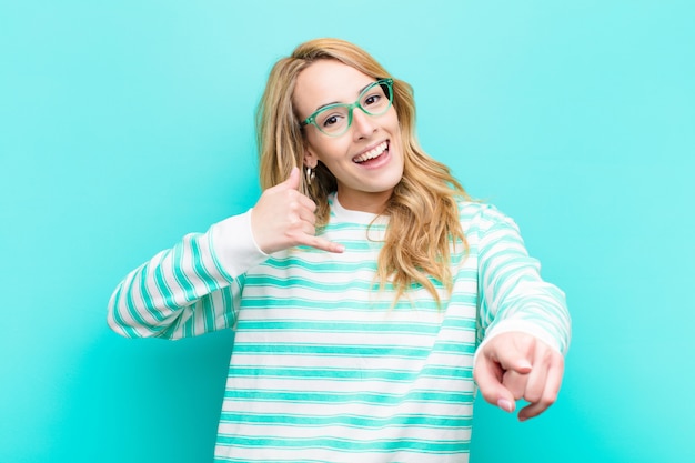 Young pretty blonde woman smiling cheerfully and pointing to camera while making a call you later gesture, talking on phone against flat color wall