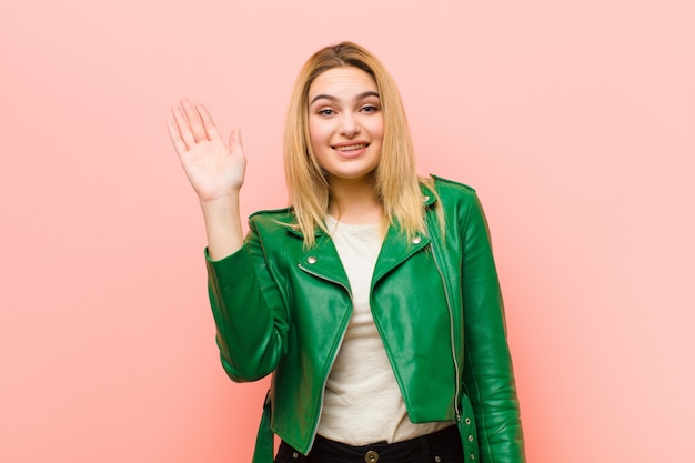 Giovane bella donna bionda che sorride allegramente e allegramente, agitando la mano, dandovi il benvenuto e salutandovi, o salutandovi sopra il muro rosa