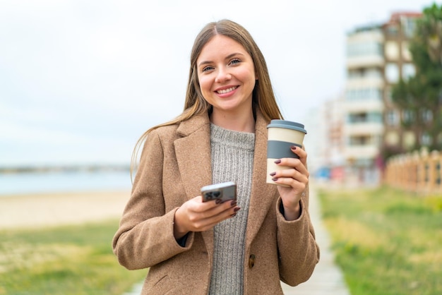 携帯電話を使用して、幸せそうな表情でコーヒーを保持している若いきれいなブロンドの女性