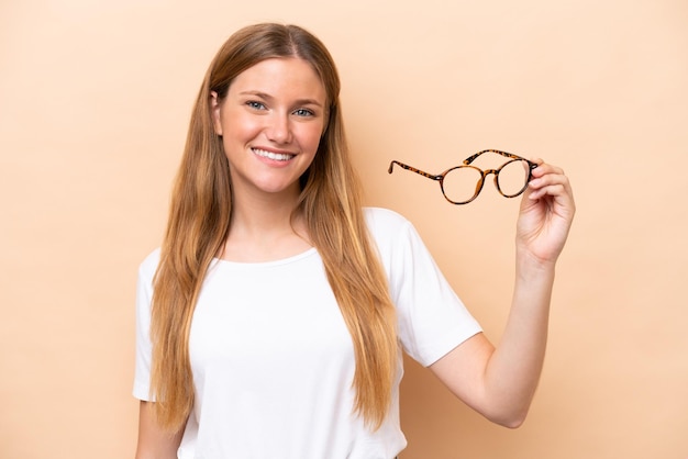 Young pretty blonde woman with glasses isolated on beige background smiling a lot