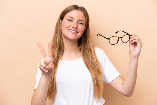 Young pretty blonde woman with glasses isolated on beige background smiling and showing victory sign