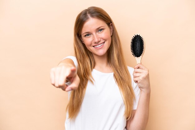 Young pretty blonde woman with hair comb isolated on beige background points finger at you with a confident expression