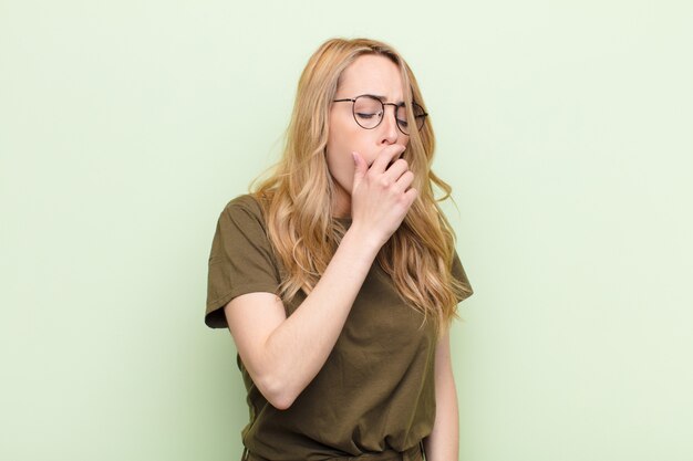 Young pretty blonde woman yawning lazily early in the morning, waking and looking sleepy, tired and bored against flat color wall