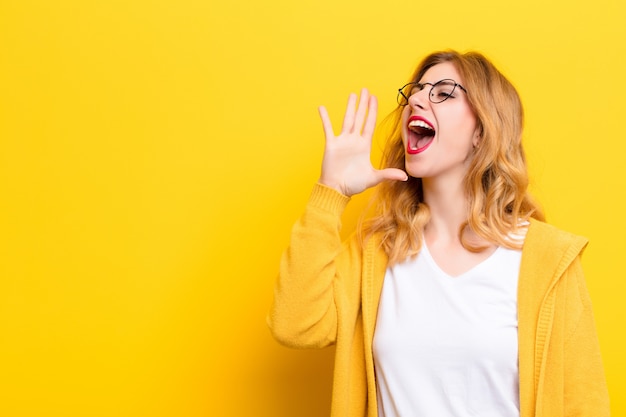 Young pretty blonde woman yelling loudly and angrily on the side, with hand next to mouth over yellow wall