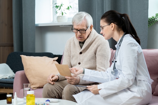 Young pretty brunette general practitioner pointing at paper with her recommendations while consulting sick senior man in living-room