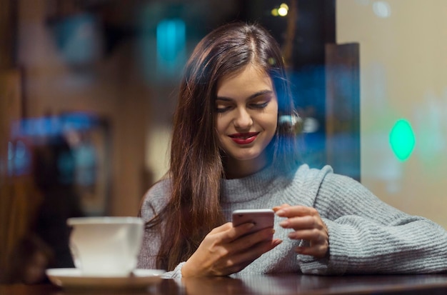 Молодая симпатичная брюнетка в сером свитере просматривает интернет через смартфон, сидя у окна кафе