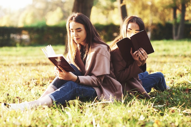 Young pretty brunette twin sisters sitting back to back on the grass with legs slightly bent in knees with brown books in hands, wearing casual coat in autumn sunny park on blurry background.