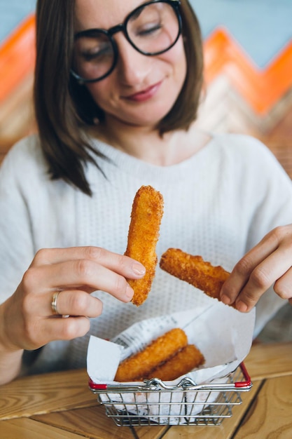 写真 若いかなりブルネットの女性は、カフェで飾られた鉄のバスケットから揚げチーズスティックを食べます。ファストフード。調色。