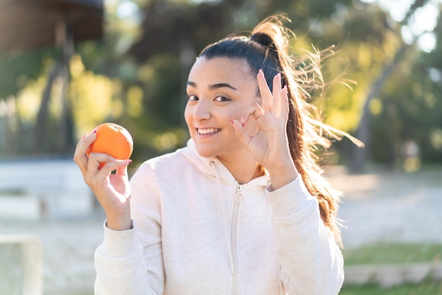 写真 指で ok サインを示す屋外でオレンジを保持している若いかなりブルネットの女性