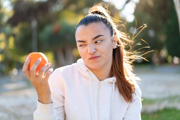 Young pretty brunette woman holding an orange at outdoors with sad expression