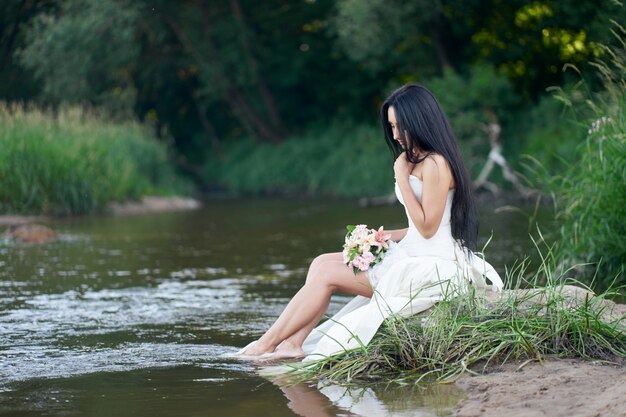 Young pretty brunette woman in white wedding dress sitting on the rock near forest river