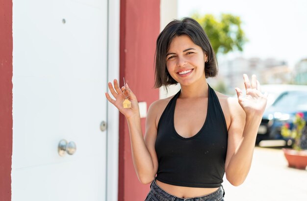 Photo young pretty bulgarian woman holding home keys at outdoors saluting with hand with happy expression