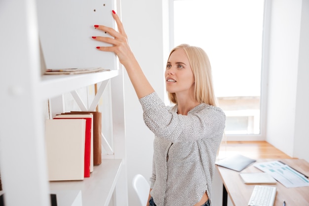 Young pretty business woman taking book from a shelf while standing in office