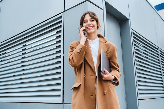 Young pretty businesswoman in coat with laptop joyfully looking in camera while talking on cellphone outdoor