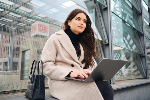 Young pretty businesswoman dreamily working on laptop on city street