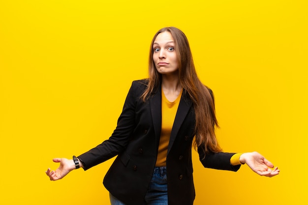 Young pretty businesswoman feeling clueless and confused, having no idea, absolutely puzzled with a dumb or foolish look against orange background