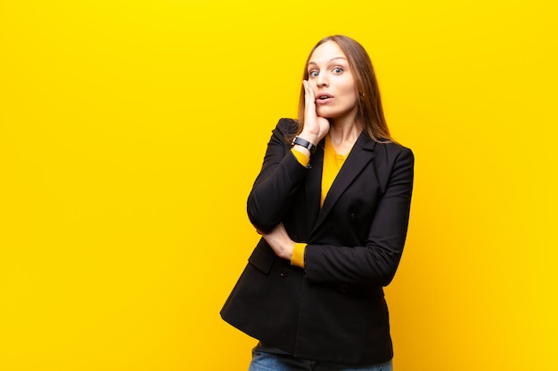 Young pretty businesswoman feeling shocked and astonished holding face to hand in disbelief with mouth wide open against orange 