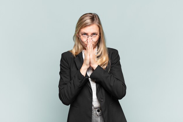 Young pretty businesswoman feeling worried, hopeful and religious, praying faithfully with palms pressed, begging forgiveness