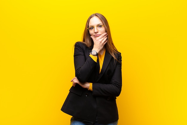 Young pretty businesswoman looking serious, thoughtful and distrustful, with one arm crossed and hand on chin, weighting options against orange wall