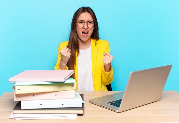 Young pretty businesswoman shouting aggressively with annoyed, frustrated, angry look and tight fists, feeling furious