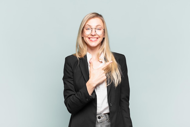 Young pretty businesswoman smiling cheerfully, feeling happy and pointing to the side and upwards, showing object in copy space