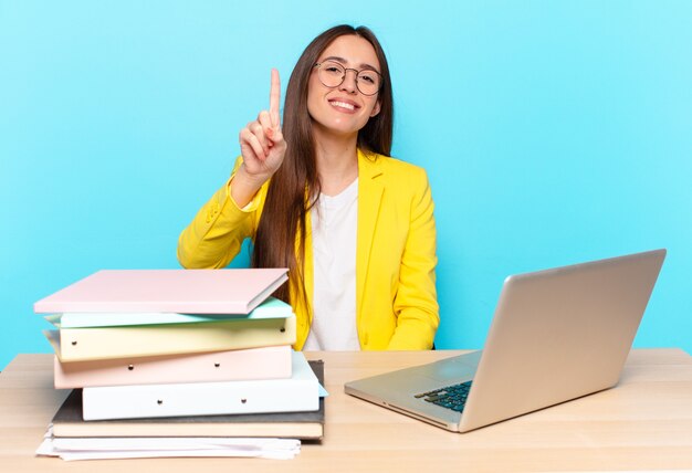 Young pretty businesswoman smiling and looking friendly, showing number one or first with hand forward, counting down