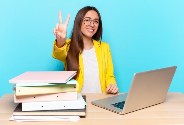 Young pretty businesswoman smiling and looking happy, carefree and positive, gesturing victory or peace with one hand